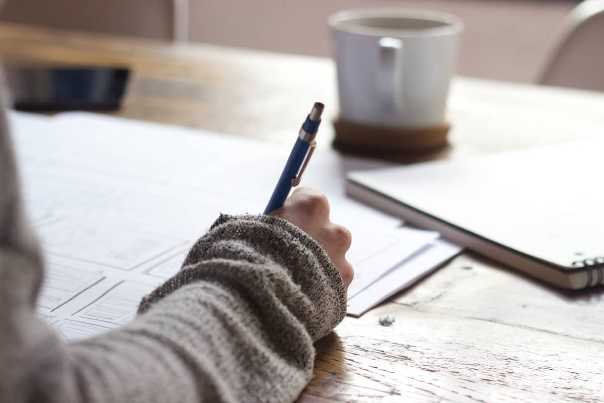 Person writing plans on paper at a wooden table beside a coffee mug and notebook