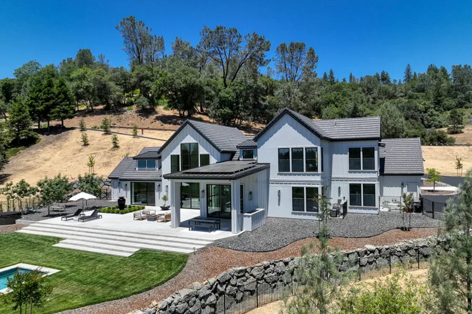 Aerial view of a new white board-and-batten custom home with gravel and rock-wall defensible space on a dry foothill hillside
