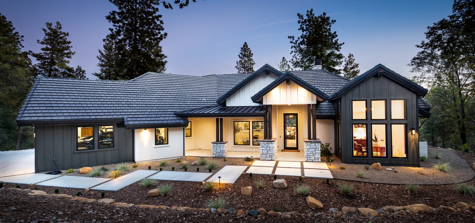 Board-and-batten custom home with stone-based porch columns and warmly lit windows at twilight beneath tall pines in Meadow Vista